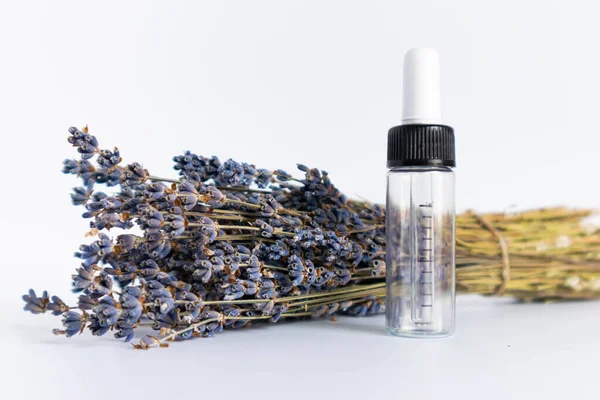 Beyaz arka planda kurutulmuş lavanta çiçekleri. Lavanta yağı. Pipetli şeffaf bir cam şişe buketin yanında duruyor..