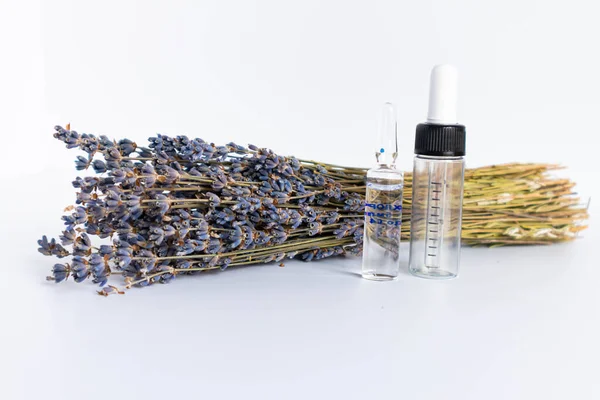 Beyaz arka planda kurutulmuş lavanta çiçekleri. Lavanta yağı. Pipetli şeffaf bir cam şişe buketin yanında duruyor..