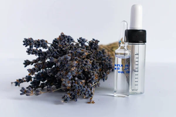 Beyaz arka planda kurutulmuş lavanta çiçekleri. Lavanta yağı. Pipetli şeffaf bir cam şişe buketin yanında duruyor..