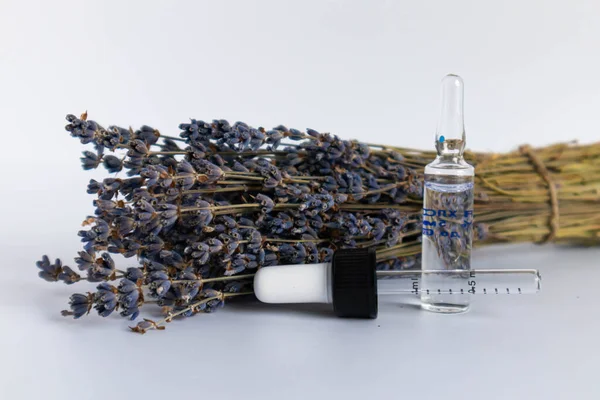 Beyaz arka planda kurutulmuş lavanta çiçekleri. Lavanta yağı. Pipetli şeffaf bir cam şişe buketin yanında duruyor..