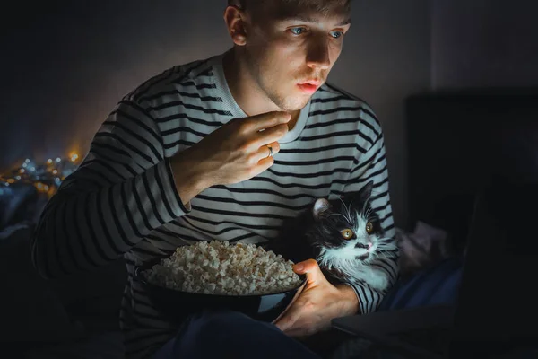 young man with black cat watching a movie eating popcorn on TV at home. Movie night. Relax,rest watching a horror film or video on screen. Background lighting. Fun Scared excited people on the couch.