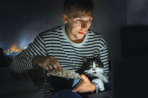 young man with black cat watching a movie eating popcorn on TV at home. Movie night. Relax,rest watching a horror film or video on screen. Background lighting. Fun Scared excited people on the couch.