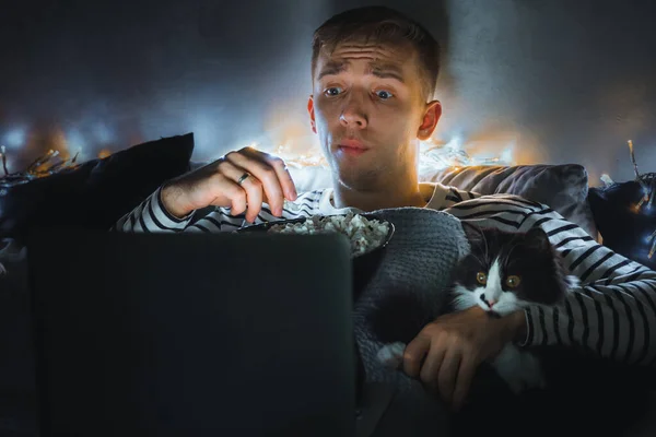 young man with black cat watching a movie eating popcorn on TV at home. Movie night. Relax,rest watching a horror film or video on screen. Background lighting. Fun Scared excited people on the couch.