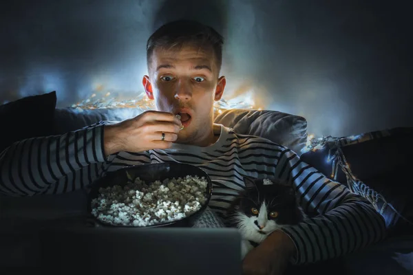 young man with black cat watching a movie eating popcorn on TV at home. Movie night. Relax,rest watching a horror film or video on screen. Background lighting. Fun Scared excited people on the couch.