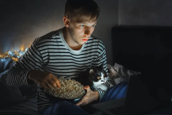 young man with black cat watching a movie eating popcorn on TV at home. Movie night. Relax,rest watching a horror film or video on screen. Background lighting. Fun Scared excited people on the couch.