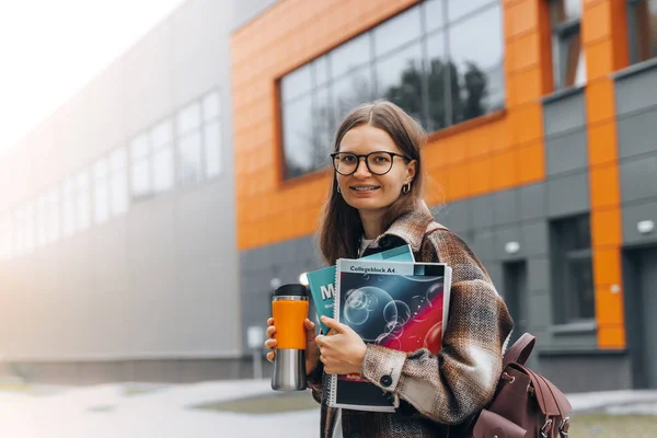 portrait of young happy student holding books coffee smiling after passing exams. Smart woman on university campus. College life. Teenanger in braces, eyeglasses with bag back to school. College life.