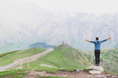 Gündelik kıyafetlerle gezen erkek turist, yeşil doğayla çevrili Gergeti Üçlemeci Kilisesi 'ne bakıyor. Kazbegi Ulusal Parkı 'nda Seyahat.
