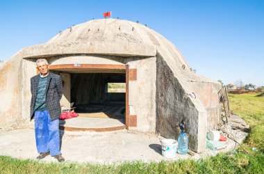 Old person stands by old italian bunker converted to home.Albania Unusual homes and living conditions. clipart