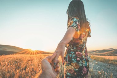Close up of a young girl in a beautiful flower dress holding hands and going towards setting sun over the horizon with golden field and scenic landscape in the background. Dramatic views and joy of travelous lifestyle concept. clipart