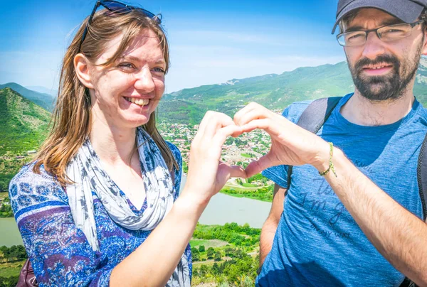Young female and male forms a heart with hands with Samtavro content church in the middle and background of caucasusu green mountains. Travel joys and religion concept
