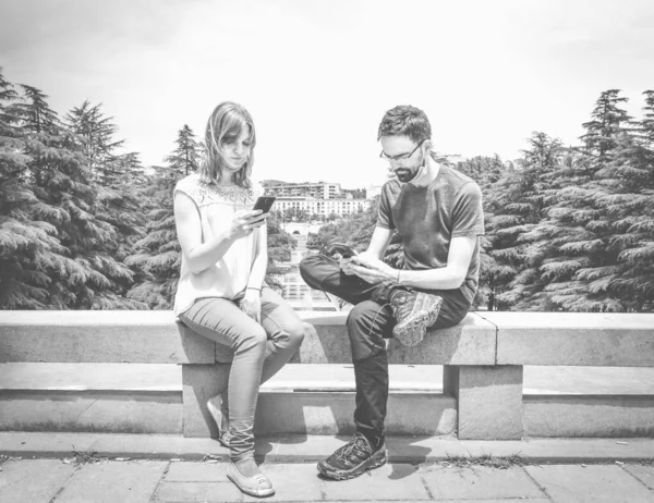 Black and white image of two young people with masks off  sitting on the cement blocks in a park and looking to their phones. Concept of ignoring social distancing and new norms in post pandemic world.