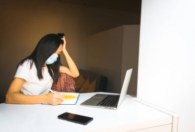 Siyah saçlı, beyaz maskeli güzel beyaz kadın rahat ev ortamındaki notlara bakıyor. Laptop ve akıllı telefon beyaz masada..