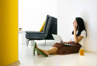 Genç beyaz kadın yerde oturuyor ve dizinde dizüstü bilgisayarla sol tarafa bakıyor..