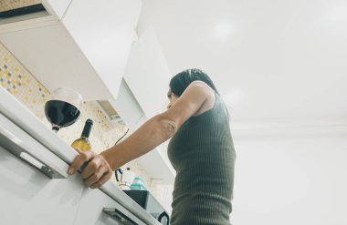 Çekici, beyaz bir kadının mutfakta dikilip şarap şişesine bir bardak şarapla baktığı düşük açılı manzara.