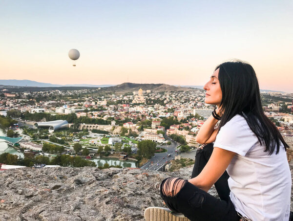 Beautiful caucasian georgian woman with closed eyes sits on the structure of narikala fortress with city view in the background.