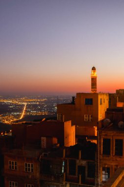 Günbatımında parıldayan şehir ışıkları ve Mezopotamya düzlükleriyle Mardin 'in eski kasabasının yukarısında aydınlanmış bir cami minaresi. Mardin Ulu Camii Minaresi Büyük Camii
