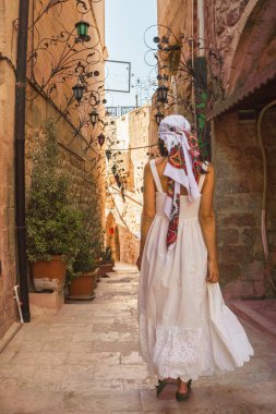 Beyaz elbiseli gezgin kadın geleneksel mimarisi ile tarihi Mardin şehrinde dar bir taş sokakta yürüyor.
