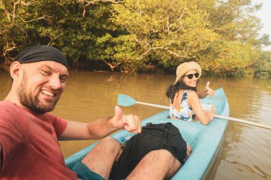 Kafkas turist çifti çift mavi kanoya oturup el sallıyor. Bereketli mangrovlar ve tropikal doğa ile çevrili sakin sularda birkaç kano.