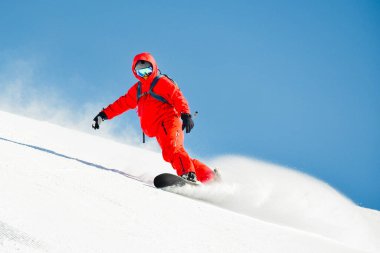 Kar kaykaycısı parlak kırmızı ceketli, yokuş aşağı kayan adam kar tozu püskürten güneşli dağ yamacına dönüyor ve açık mavi gökyüzü aşırı kış sporları yapıyor.