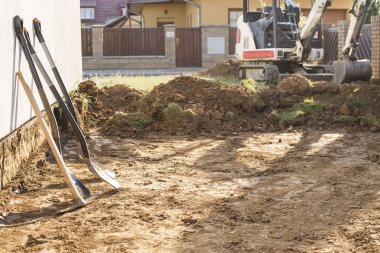 Mini Ekskavatör inşaat sitesinde. Ekskavatör evin etrafında arazi düzenlemektedir. Ön planda kürek ile toprak kazma kazıcı.