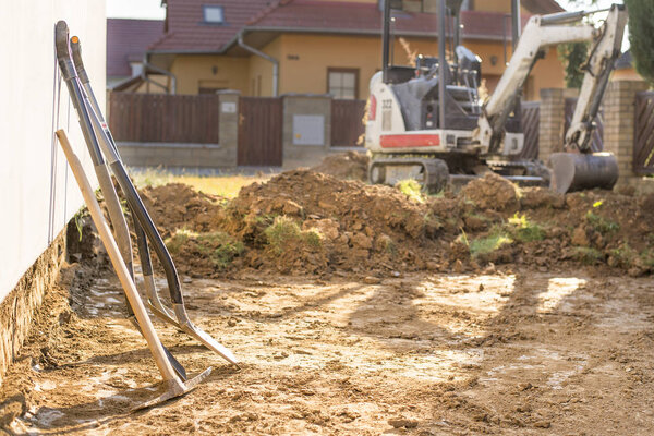 Mini excavator on construction site. Excavator regulates the terrain around the house. Digger digging soil with shovels in foreground.