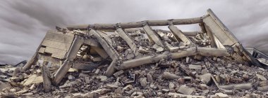 Dramatik gökyüzü ve arka planda başka bir beton bina ile çökmüş beton sanayi binası. Enkaz, toz ve hasarlı ev dolu afet sahnesi