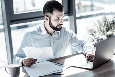 Belgeyi tutarak ve dizüstü bilgisayar kullanarak ciddi adam konsantre.