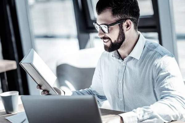 Satisfecho empleado agradable sonriendo y con vistas al cuaderno . — Foto de Stock