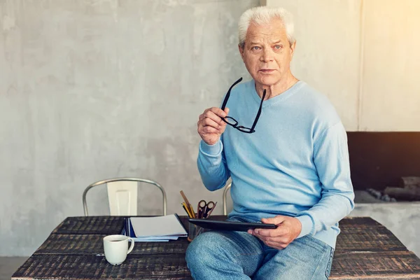 Thoughtful mood. Serious senior man holding a tablet and a pair of glasses while sitting on a table