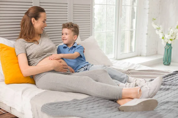 Счастливая беременная женщина лежит на двуспальной кровати с маленьким сыном — стоковое фото