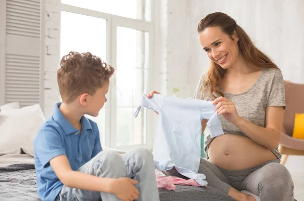 Mujer embarazada radiante mostrando ropa para su pequeño bebé a su hijo — Foto de Stock