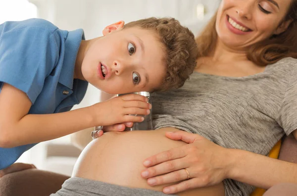 Curieux garçon aux yeux foncés écoutant le ventre enceinte — Photo