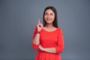 Mükemmel bir fikir. Kırmızı bir elbise onun parmak yükselterek ve izole bir mavi-gri arka plan üzerinde duran yeni bir fikir hakkında mutlu neşeli genç kadın