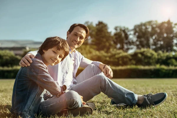 Pai e filho sentados na grama sorrindo e abraçando — Fotografia de Stock