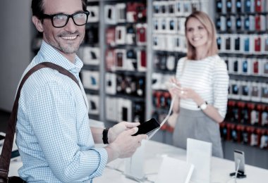 Samimi bir gülümseme. Bir akıllı telefon elektronik mağazasında olurken tutan neşeli gülümseyen kişi
