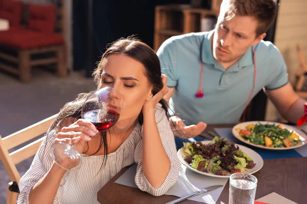 Blonde-haired man feeling angry while watching girlfriend drinking alcohol — Stock Photo, Image