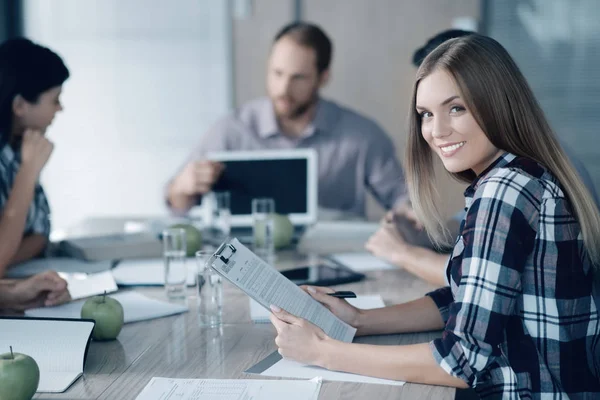 Mujer joven y positiva sentada a la mesa con sus colegas — Foto de Stock