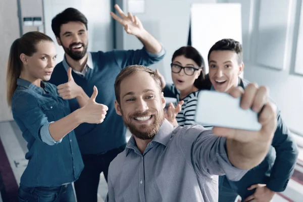 Broad smile. Cheerful bearded young man making selfies and resting with his colleagues while standing in the office