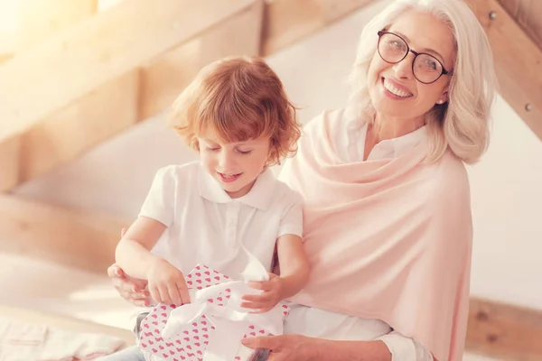 Joyful granny smiling into camera while her grandson opening gift — Stock Photo, Image