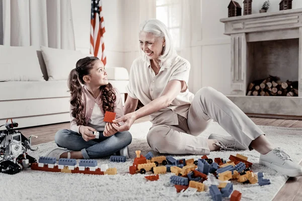 暖炉の近くの床の上に座って驚く女の子にレゴの詳細を与える笑顔素敵な晴れやかなおばあちゃん — ストック写真