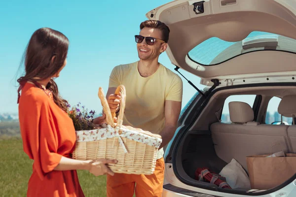 Современная счастливая пара берет корзину для пикника из своей машины — стоковое фото
