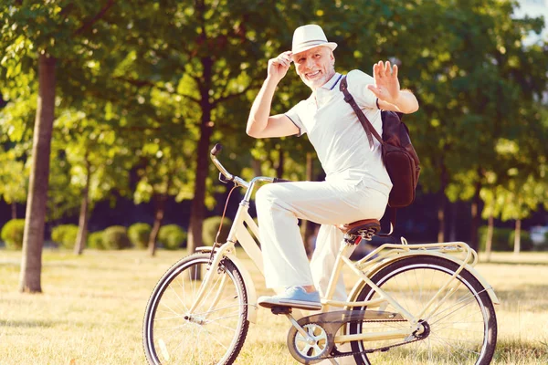 Friendly retired man sitting on bicycle and greeting somebody in park