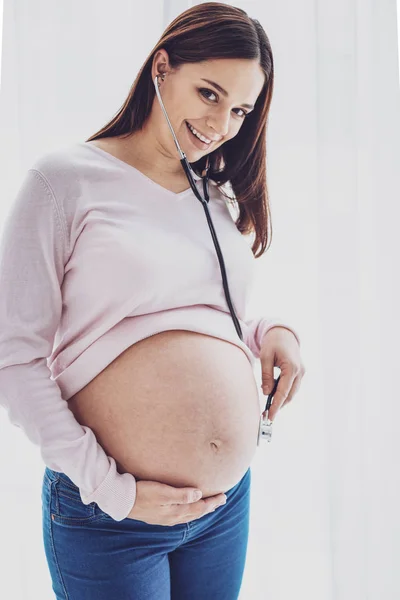 Donna allegra incinta che ascolta il suo stomaco attraverso le cuffie mediche — Foto Stock