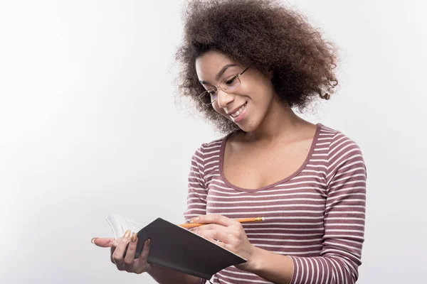 Clever dark-haired curly student doing her homework making some notes — Stock Photo, Image