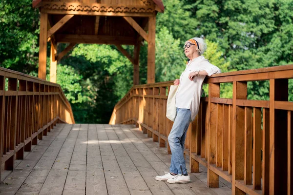 Stylish elderly woman wearing jeans and white sneakers walking in park