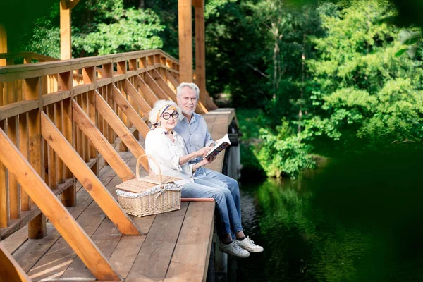 Casal idoso lendo livro interessante enquanto sentado na ponte — Fotografia de Stock