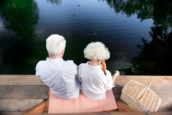 Pár důchodců sedí poblíž jejich koši krmení kachen v jezeře — Stock fotografie