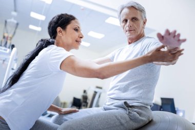 Dikkatli göz atmak. Ciddi sakin iken bir rehabilitasyon merkezinde büyük bir fitball oturan ve neşeli bir deneyimli doktor ona yardım ederken elini bakarak konsantre duygu adam yaşlı