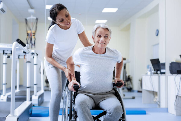 Attention. Positive emotional disabled pensioner sitting in a wheelchair and smiling cheerfully while attentive kind skilled specialist helping him