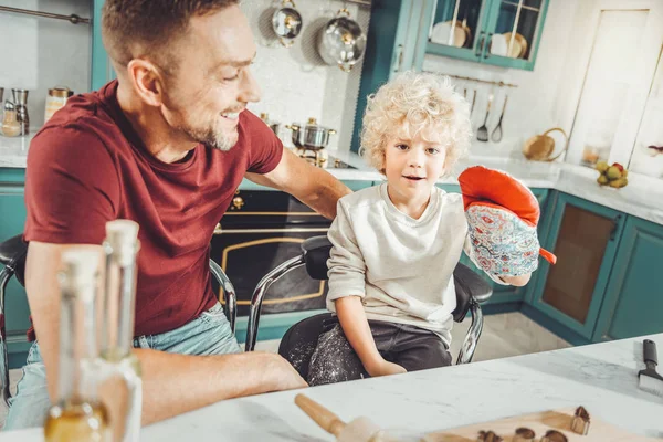 Curly blonde-haired son putting pot holder on while helping in kitchen